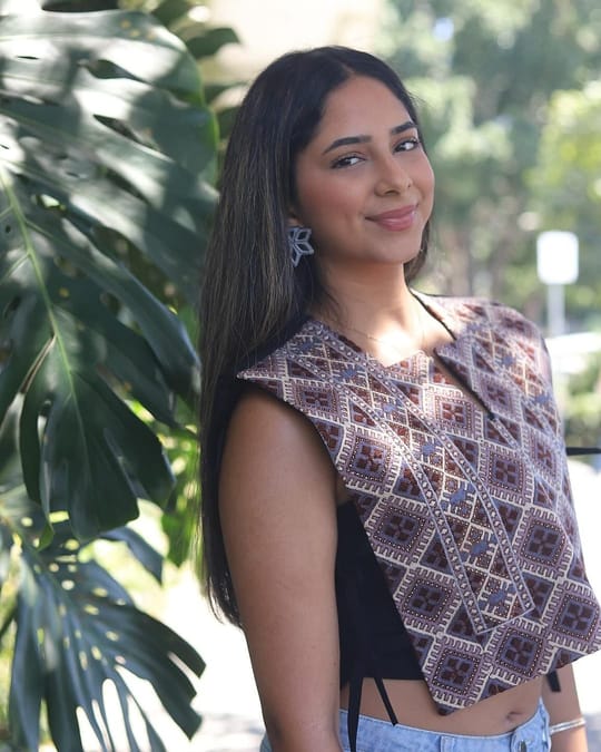 Model smiling in a cropped mauve lattice embroidered vest, beside tropical leaves