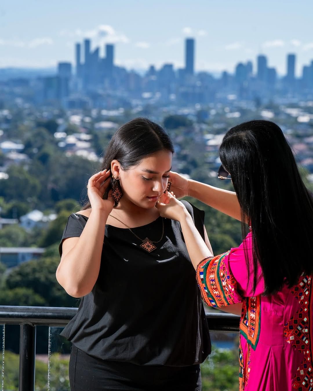 A Balochi woman in traditional dress fits handmade earrings on a model, with the Brisbane skyline behind